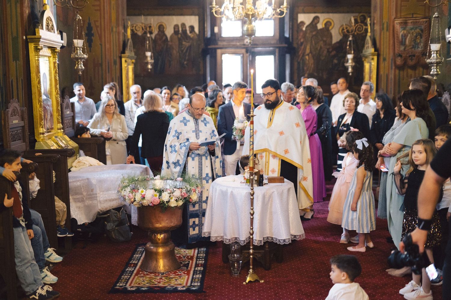 A traditional Orthodox Christian baptism ceremony taking place inside an ornate church with gold decorations and icons.