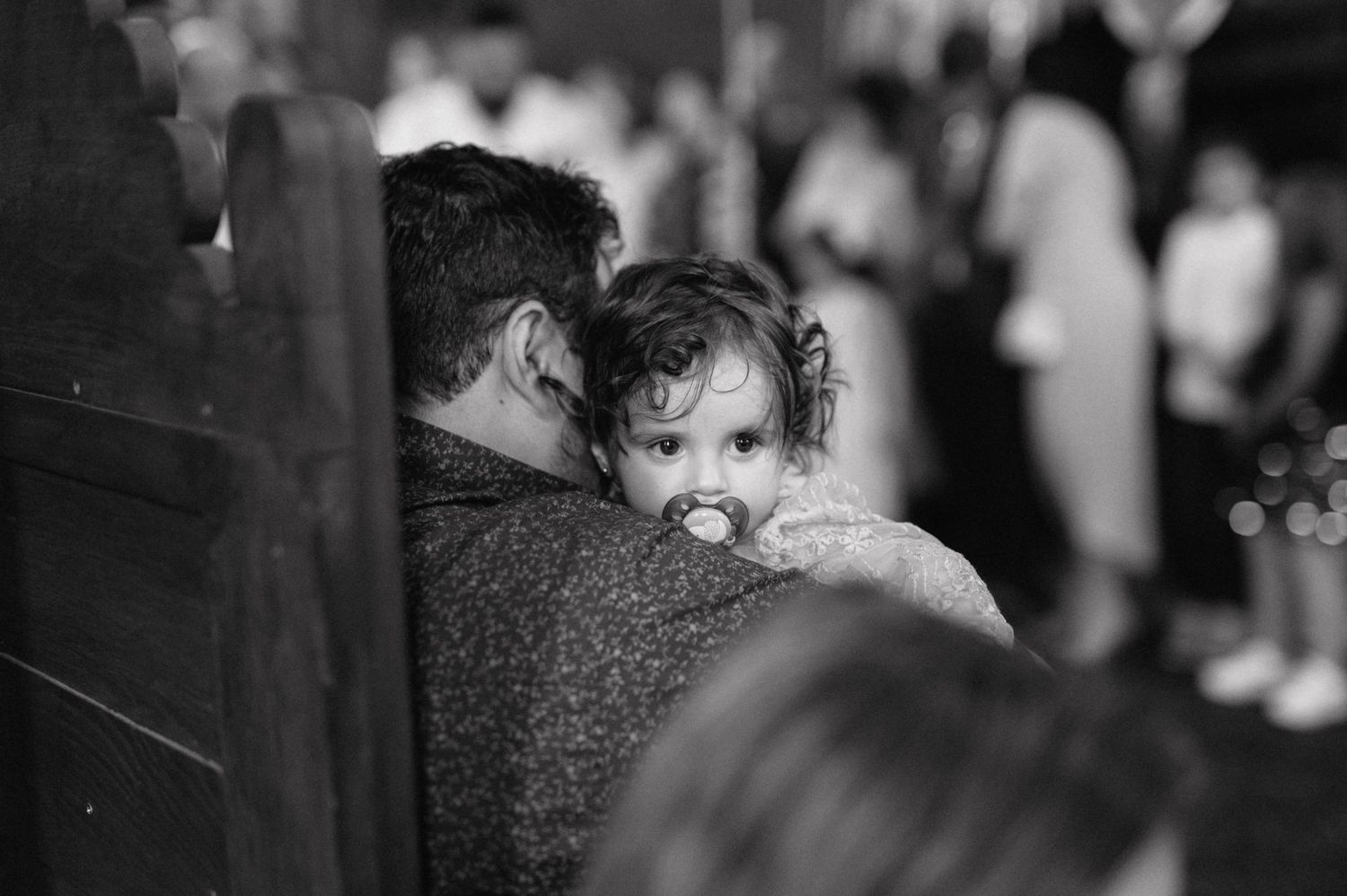 A small child with a pacifier peeks over an adult's shoulder in a black and white candid photograph.