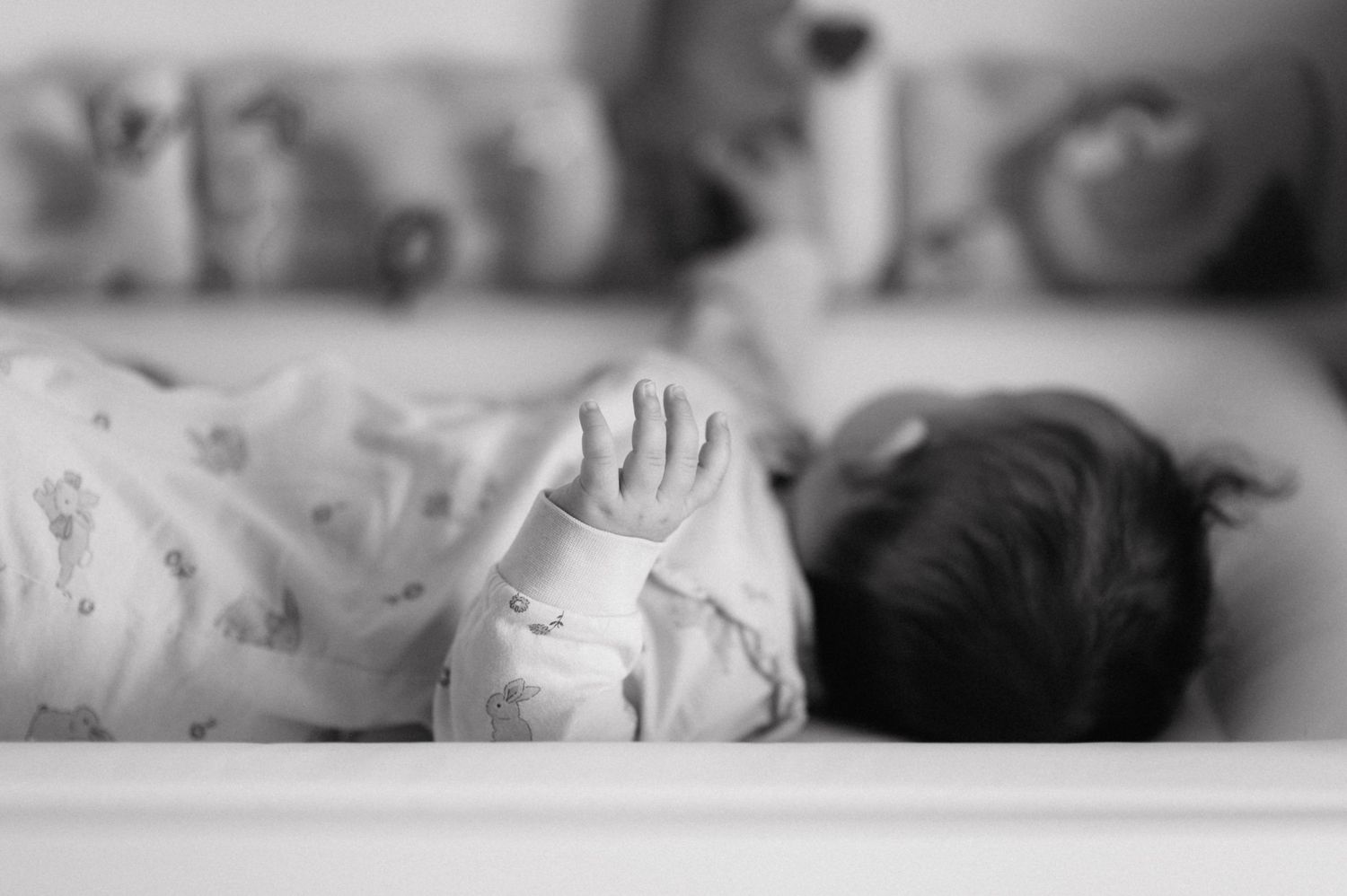 Black and white photo of a small baby lying on bedding, with a tiny hand reaching up in soft focus.