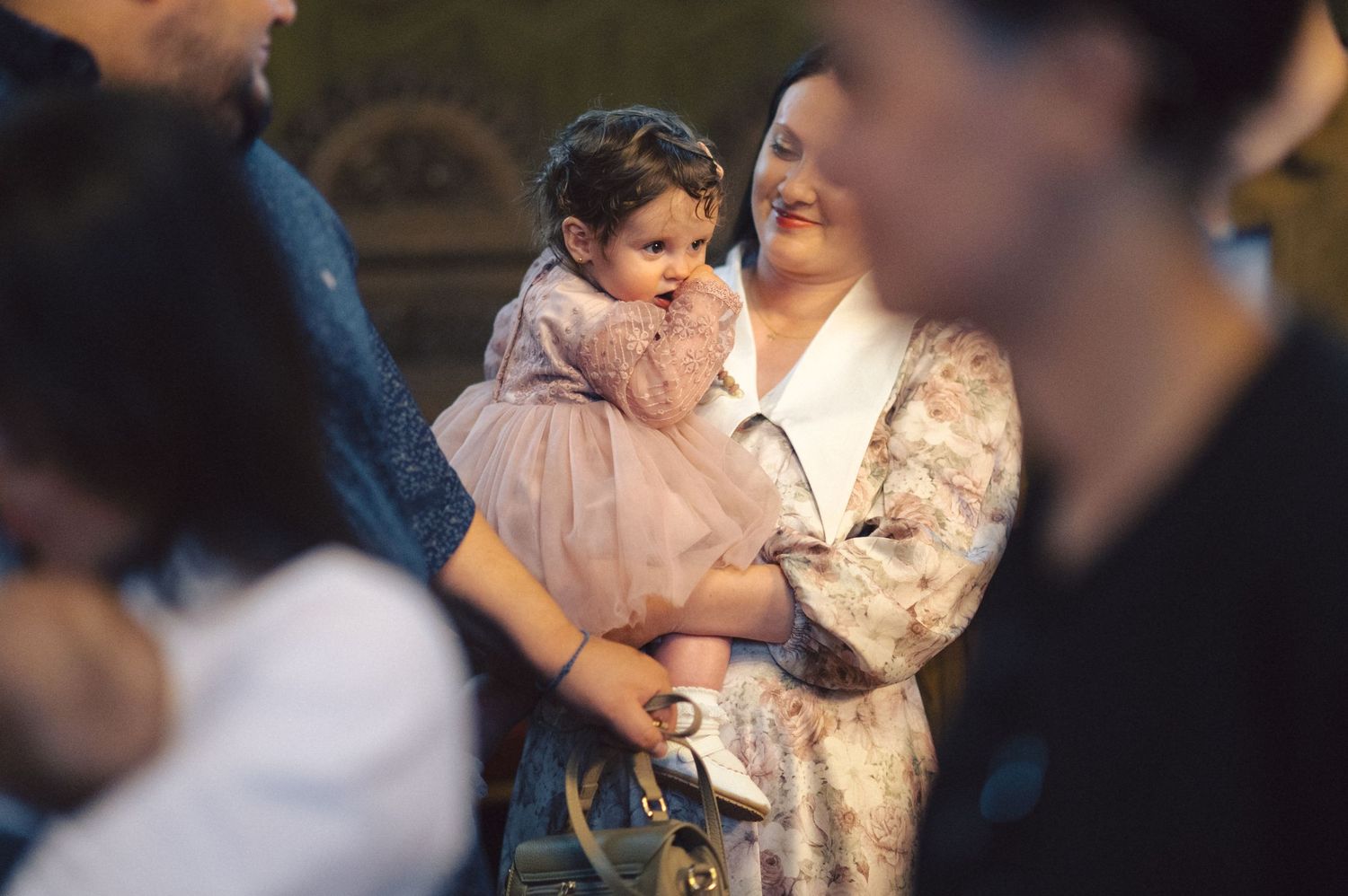 A small child in a fancy dress being held during what looks like a formal family gathering.