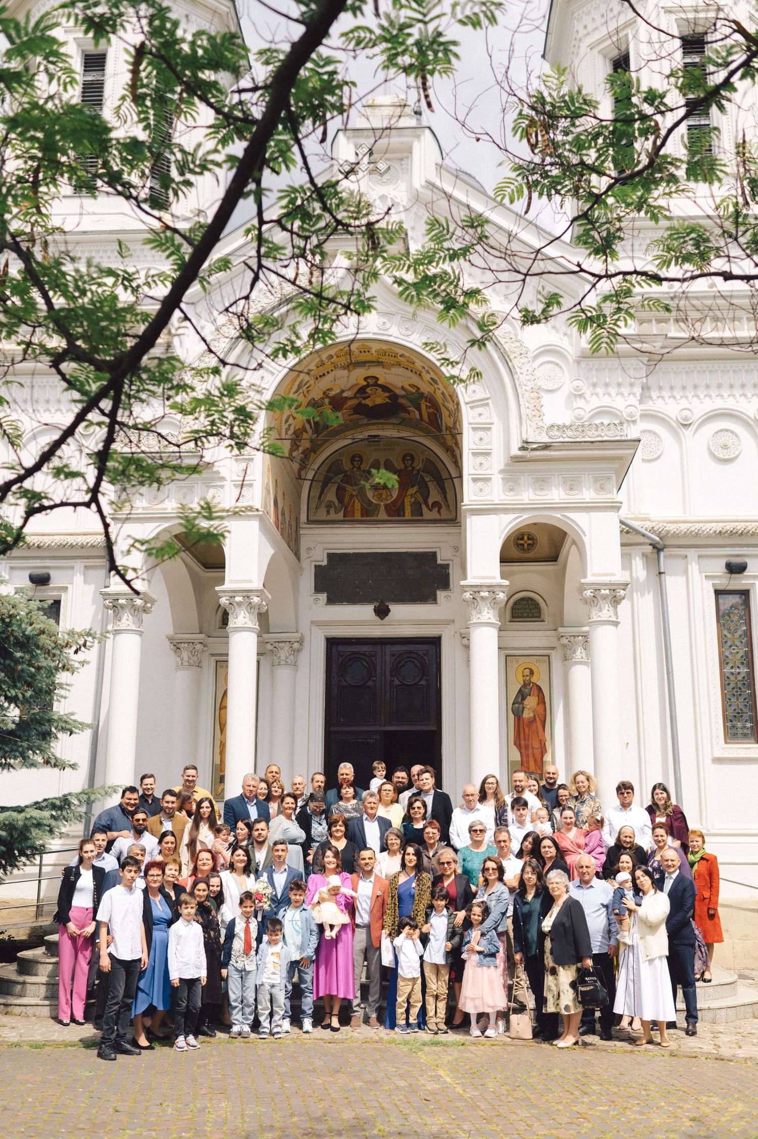 A large gathering of people posed on the steps of an ornate white church with arched doorways and detailed architecture.