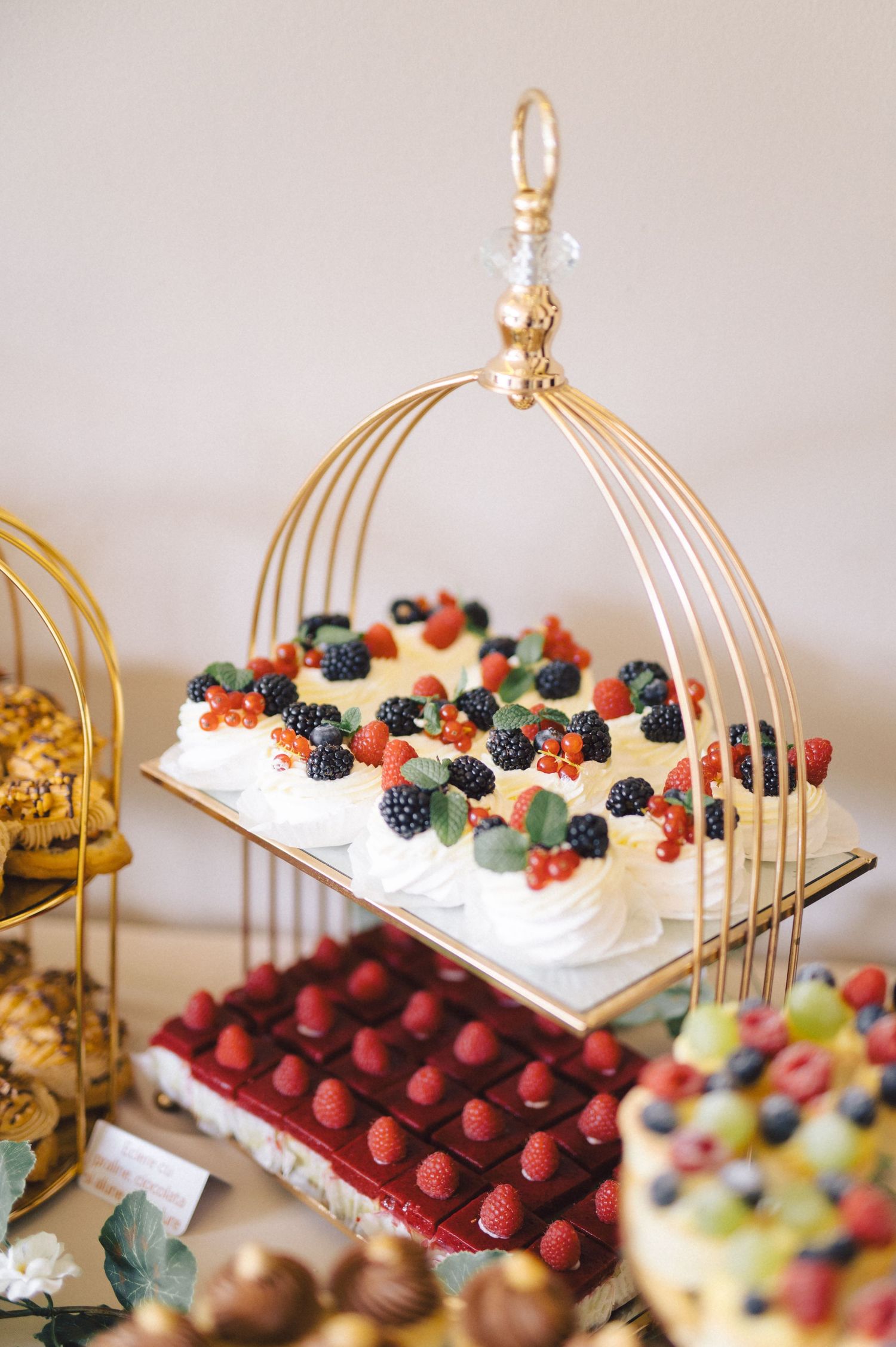 Elegant dessert display with berries atop white cream on a gold wire stand, with red cherries and other treats below.