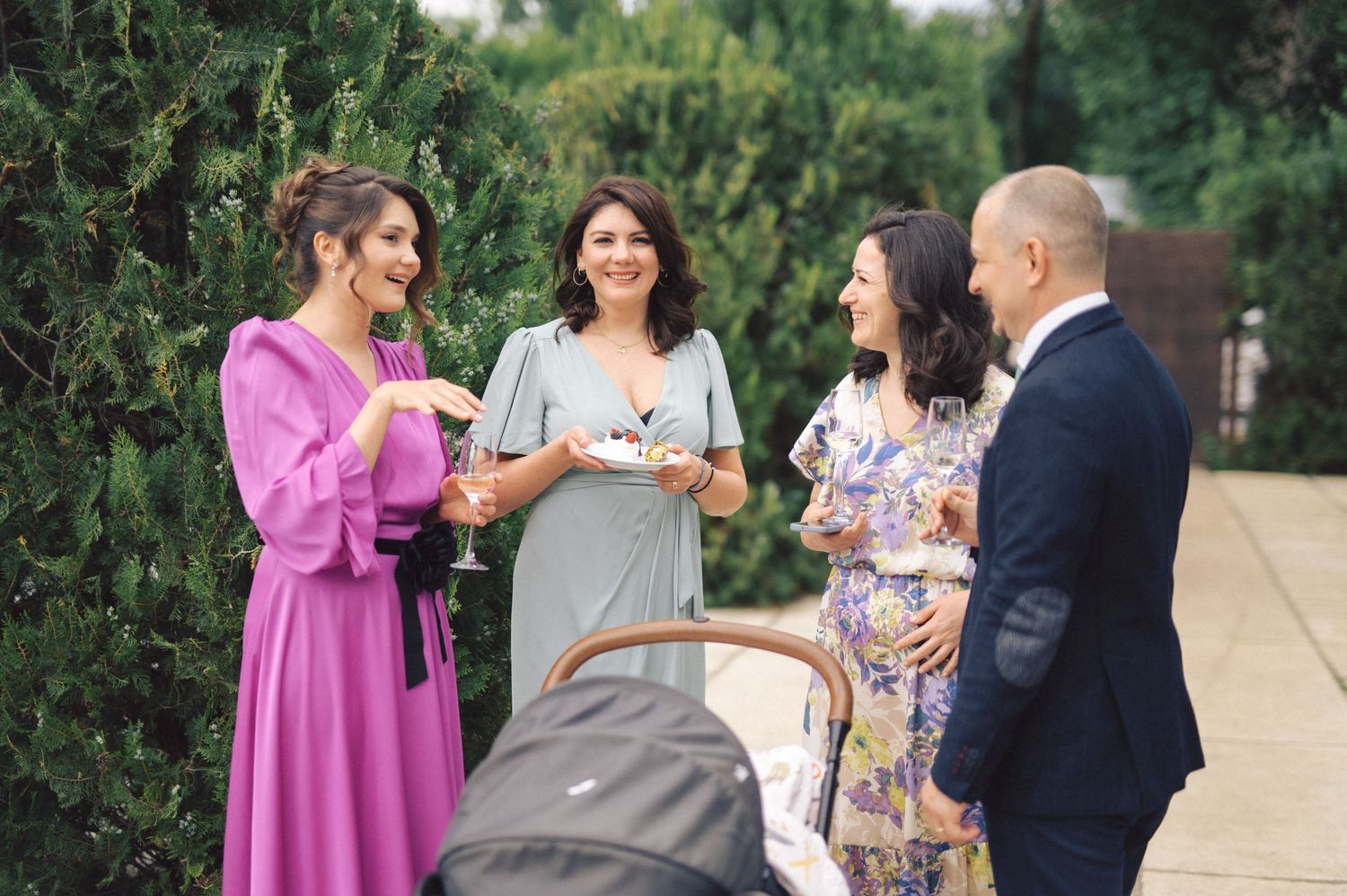 A group of people in formal attire stand together chatting outdoors next to greenery on a sunny day.