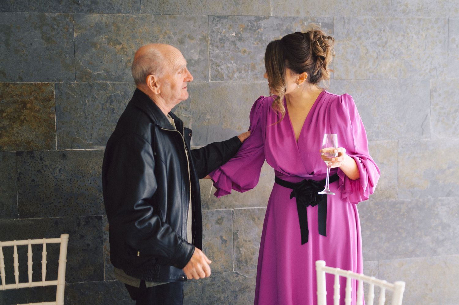 Two people chatting during an event, with one wearing a vibrant pink dress against a stone wall.