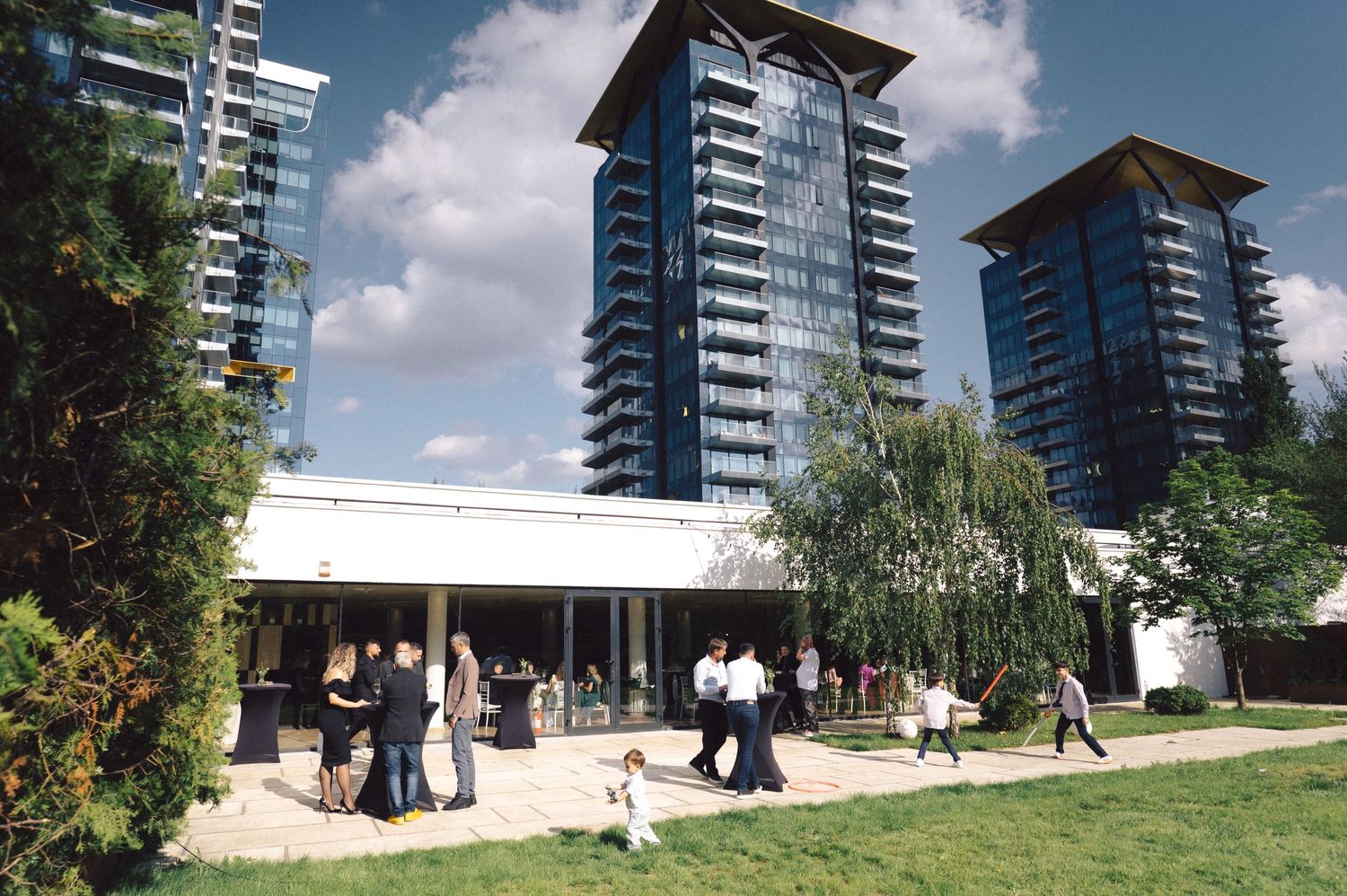 People gather outside a modern building with three glass high-rise towers in the background on a sunny day.