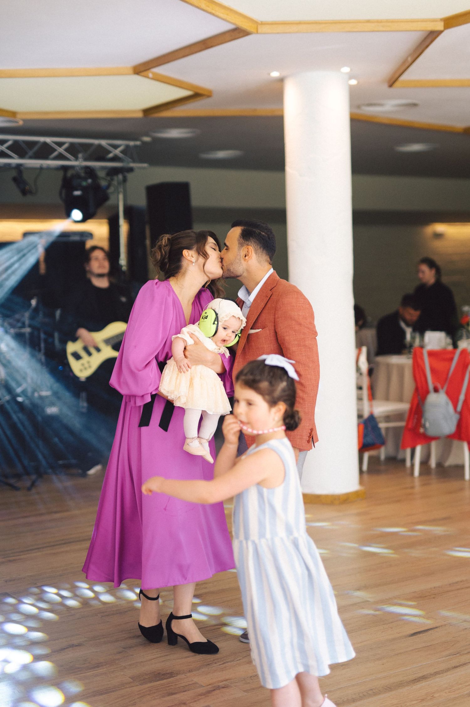 A couple shares a romantic moment on a dance floor while holding a baby doll and a child in a striped dress watches.