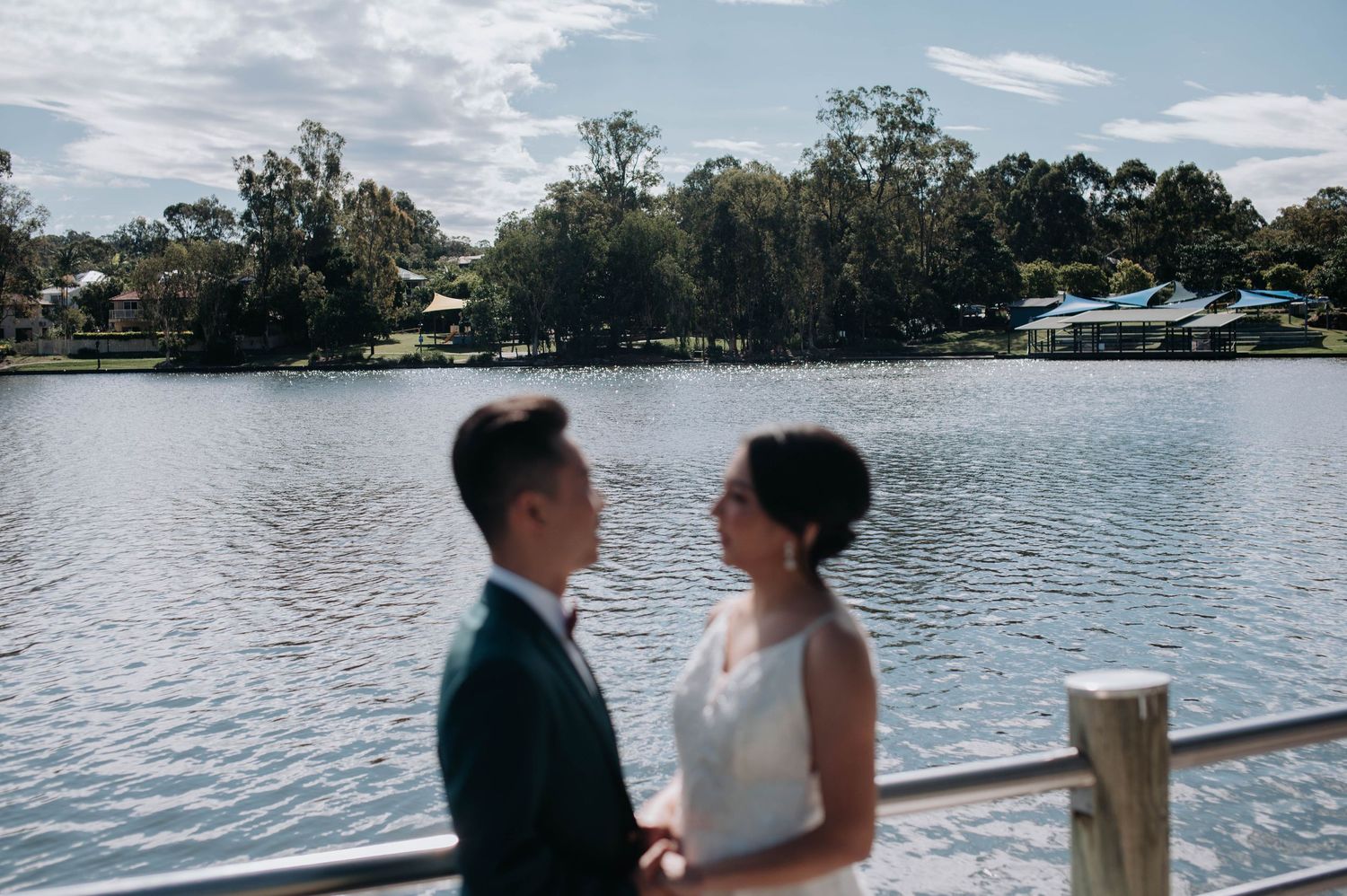 A couple in wedding attire stands together on a lakeside deck with trees and boats visible in the background.