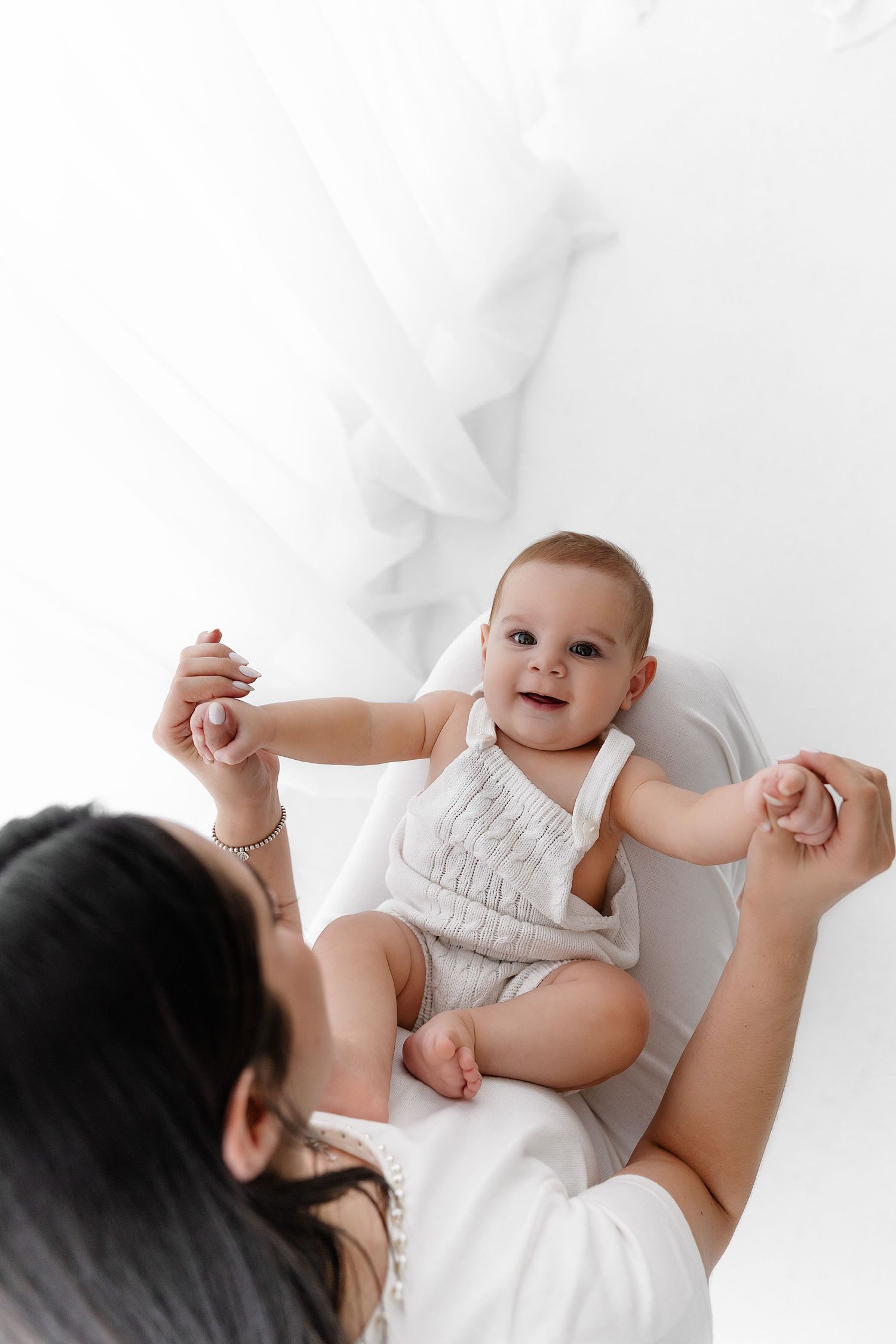 Smiling baby in white onesie lies on white blanket while being held by caregiver from above.