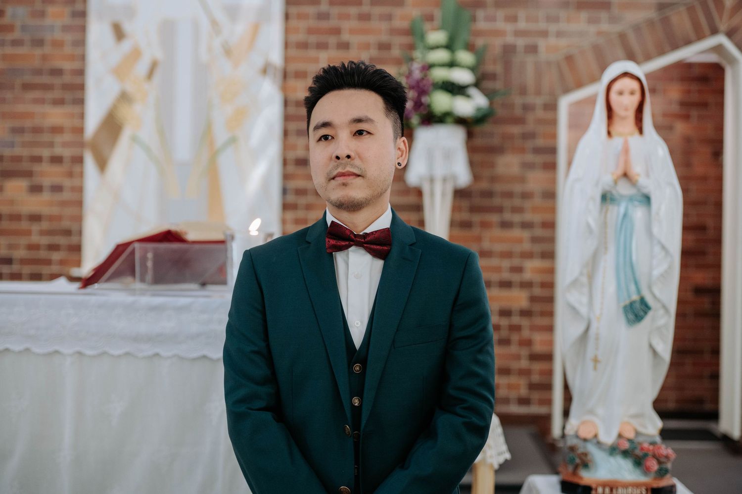 Well-dressed person in a dark green suit and bow tie stands in a church with a statue of the Virgin Mary visible.