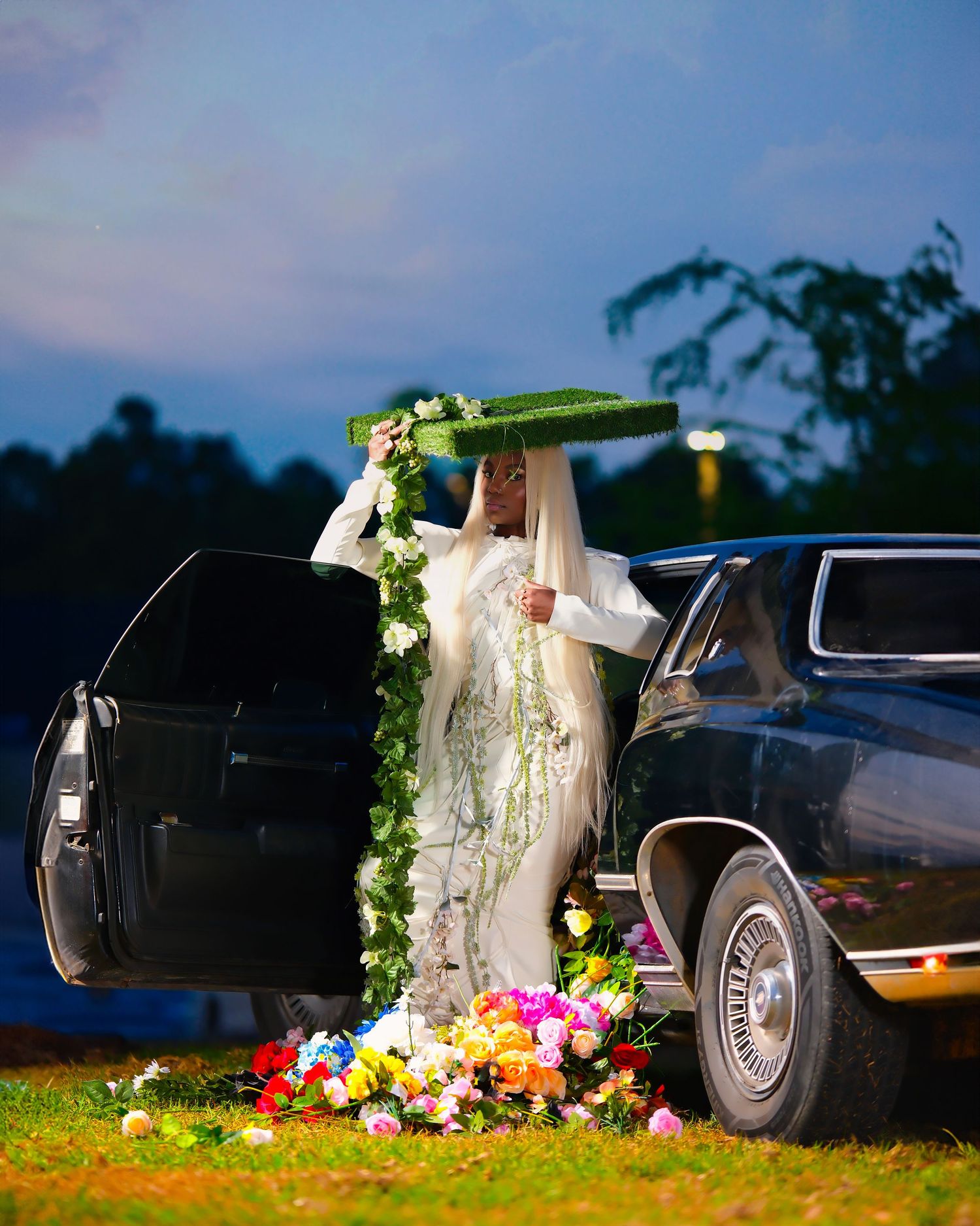 A wedding dress hangs elegantly from a decorated car door at dusk with colorful flowers scattered at the base.