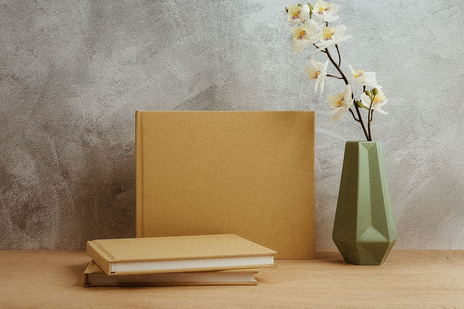 White orchid flower in modern green geometric vase beside stacked brown books on wooden surface against textured wall.