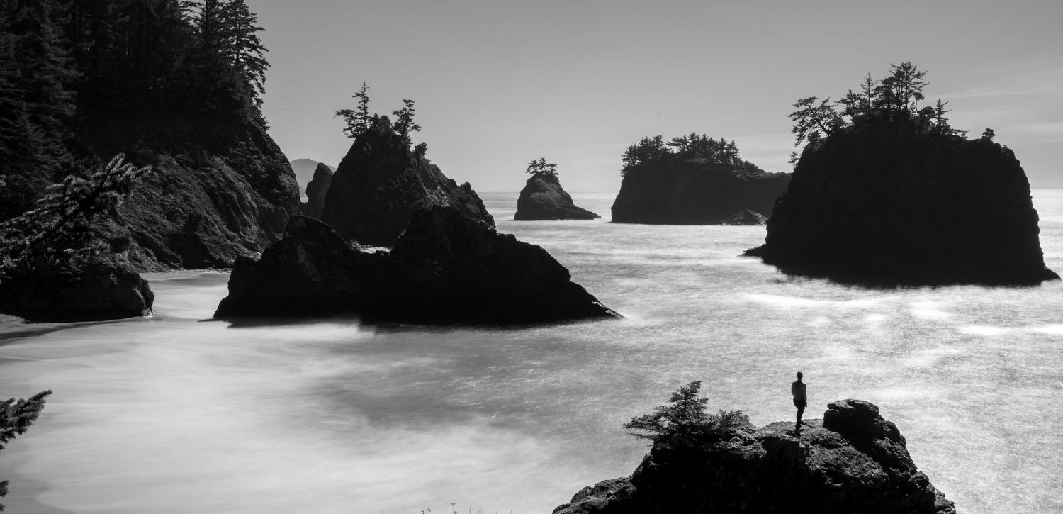 Black and white landscape of sea stacks along the Oregon coast, with textured waves and rocky shoreline under dramatic light.