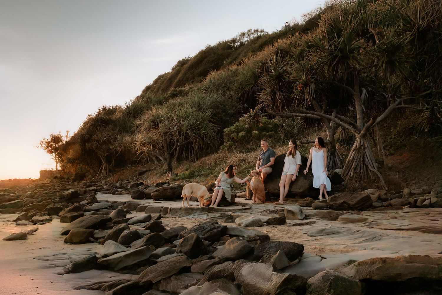People in light colored dresses gather on a rocky beach at sunset with cliffs and trees in the background.