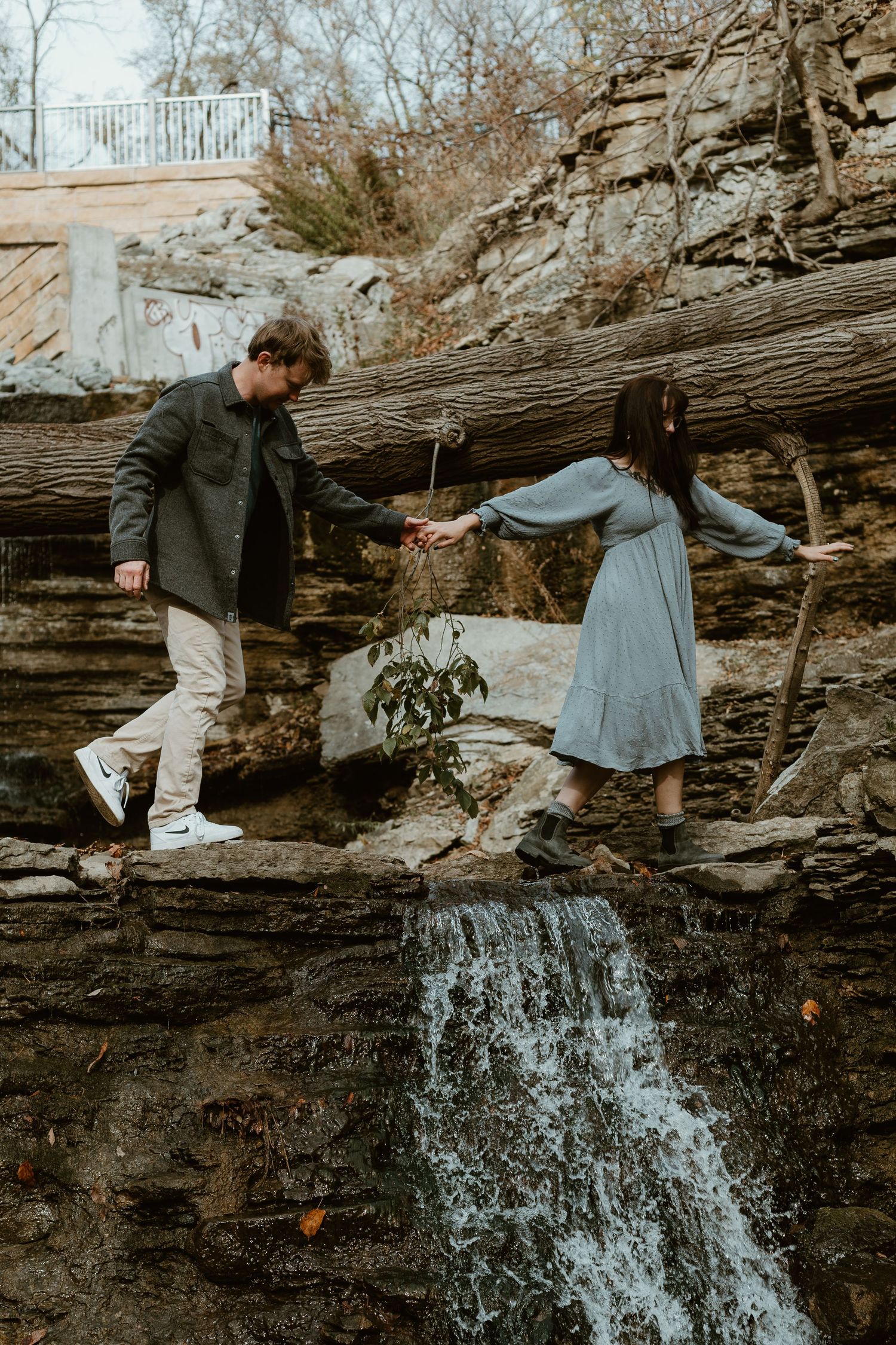 Two people walking carefully across rocky ledge near small waterfall in autumn wearing casual clothing.