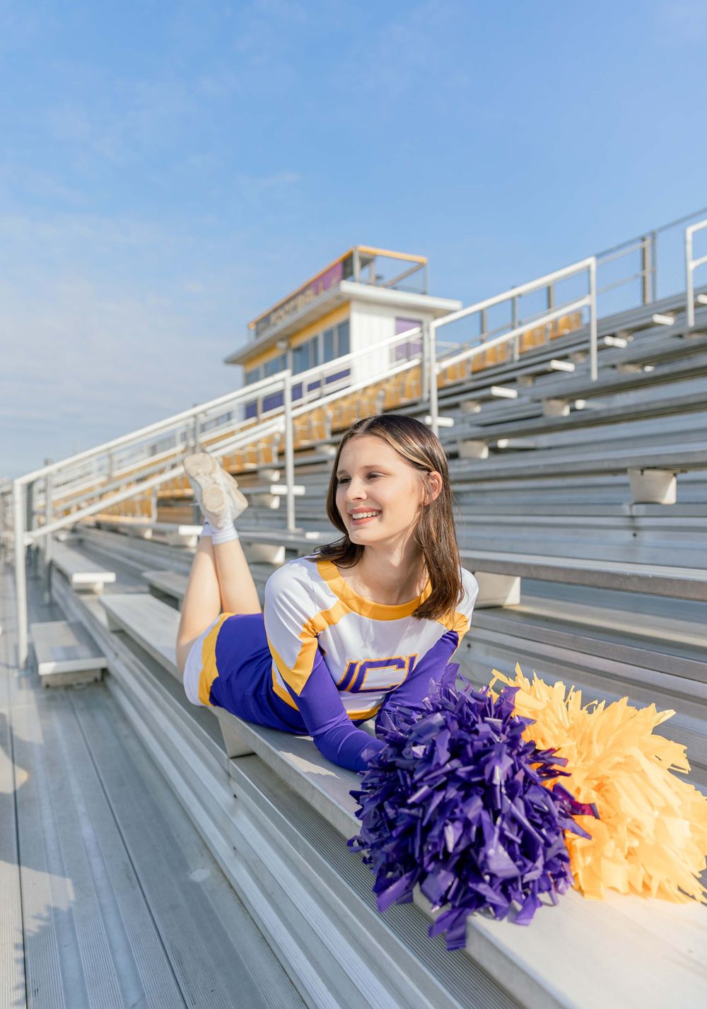 Cheerleader in purple and gold uniform poses with pom poms on stadium bleachers.
