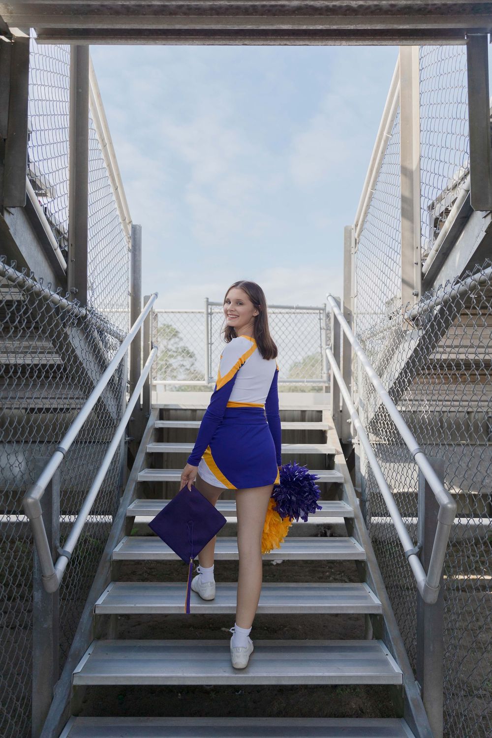 Cheerleader in blue and yellow uniform poses on metal stadium stairs with pom poms.