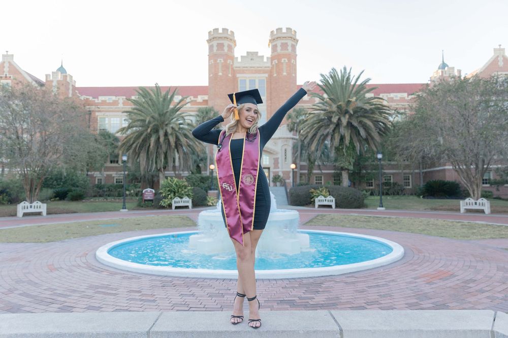 Graduate in academic regalia poses with one leg raised in front of a fountain at Florida State University campus.