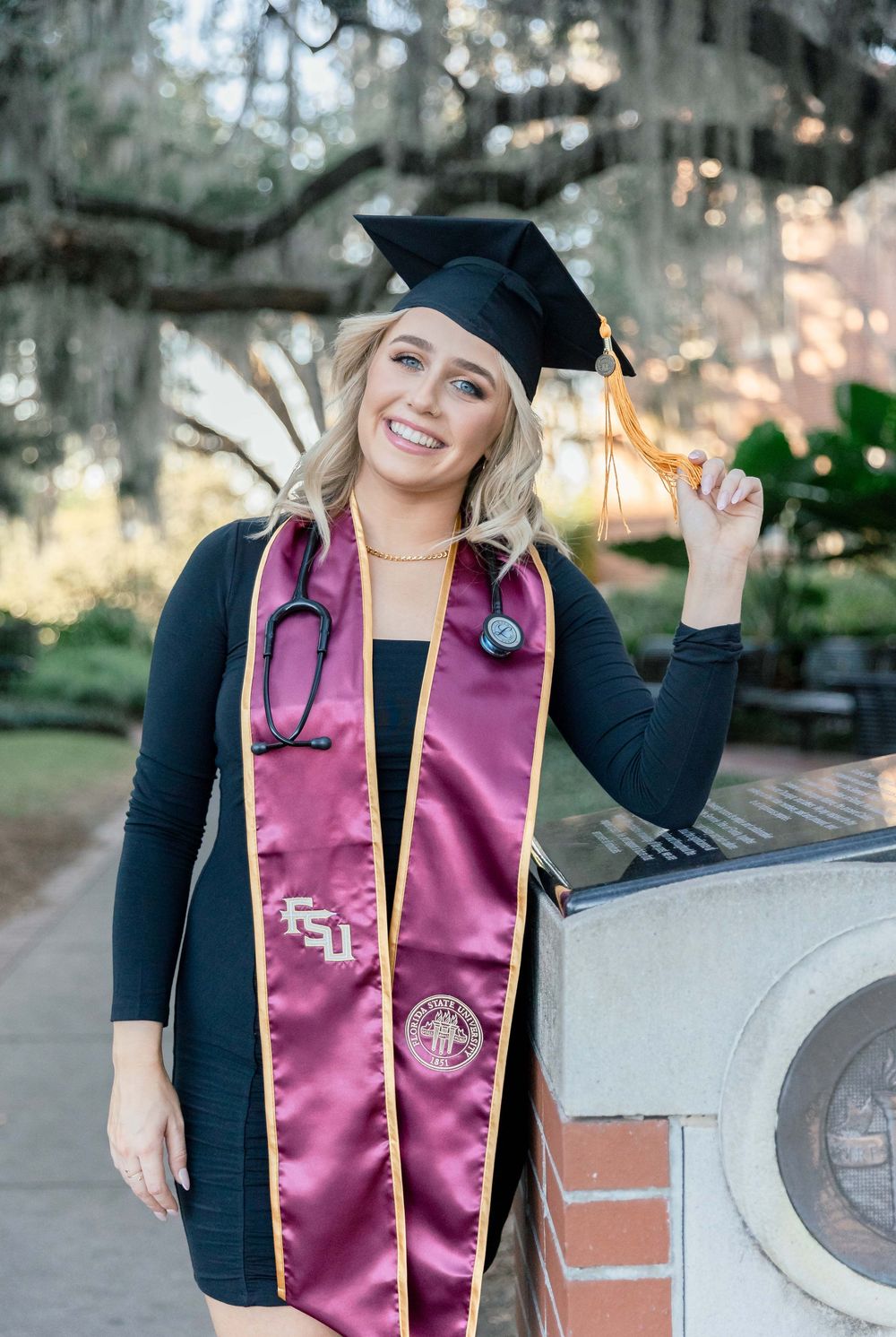 Graduate wearing black cap and gown with pink medical stethoscope and FSU sash poses outdoors during graduation photoshoot.