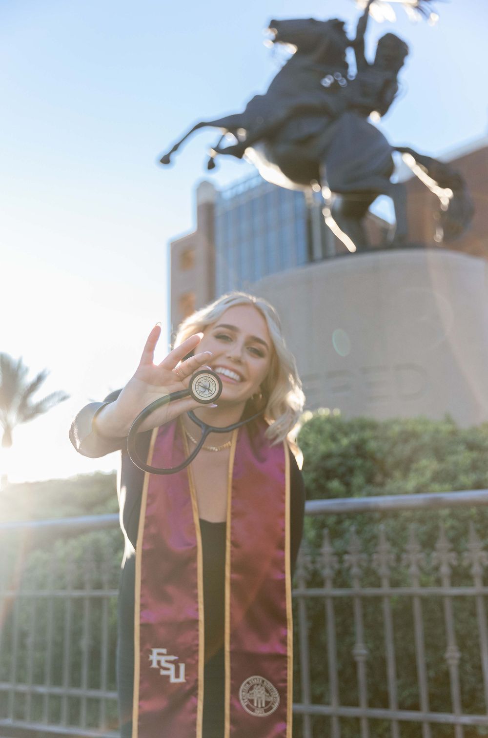 Graduate in burgundy Florida State University stole celebrates in front of campus statue during golden hour sunlight.