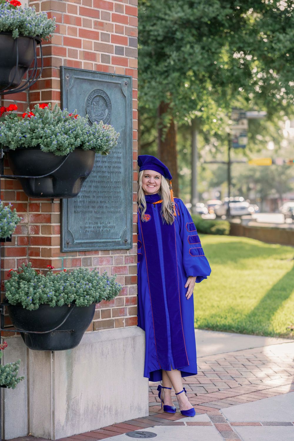 Graduate in vibrant blue cap and gown poses outdoors near brick wall with planters during commencement photo session.