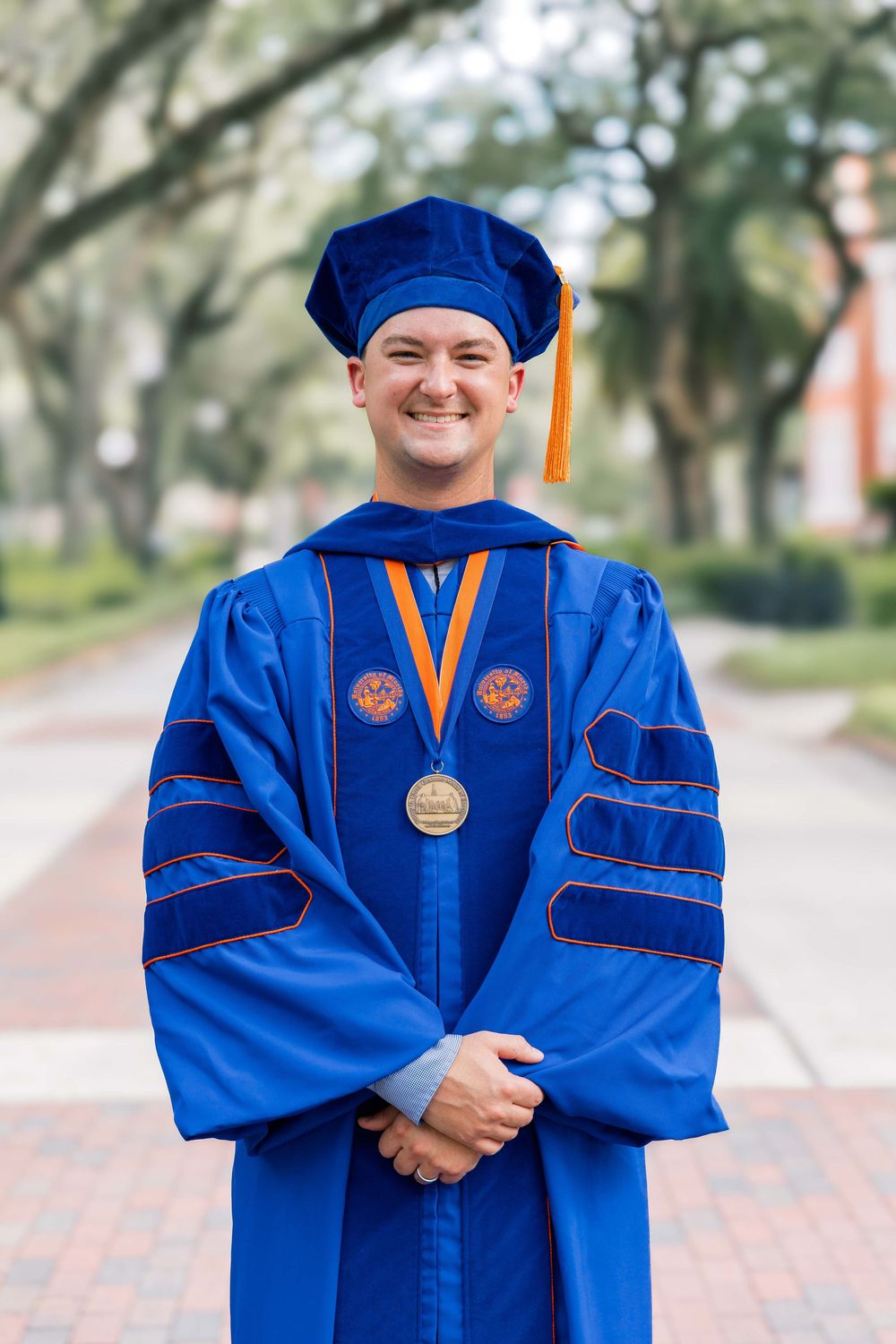 Graduate in blue doctoral regalia with orange accents stands proudly on campus path.
