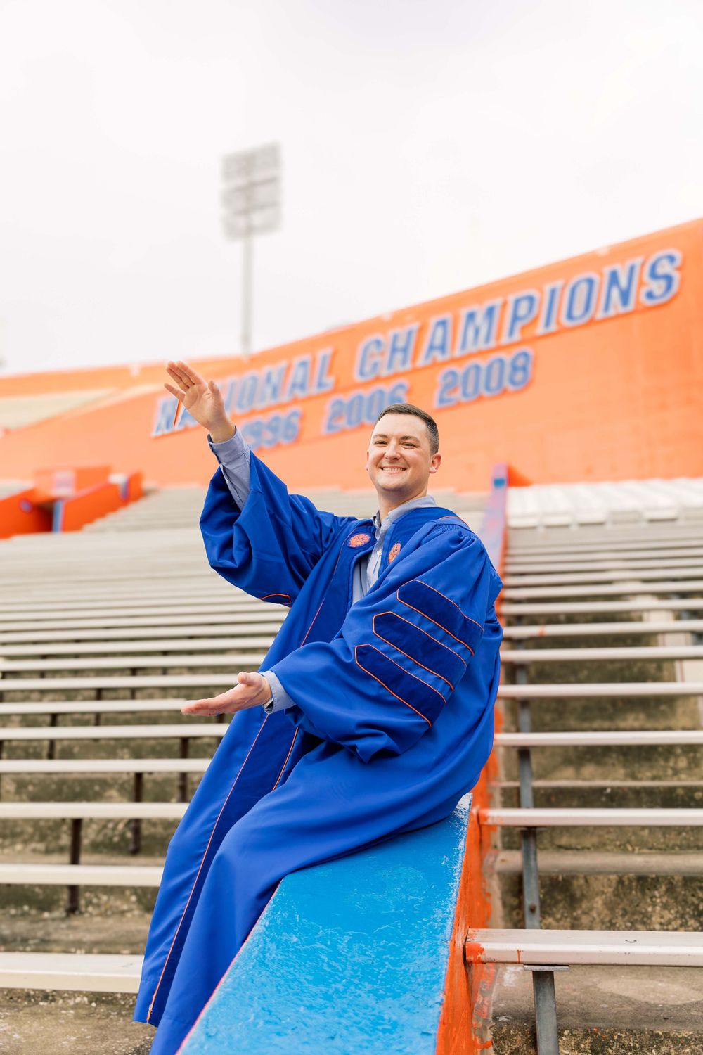 Graduate in blue academic regalia poses joyfully on stadium bleachers near championship signage.