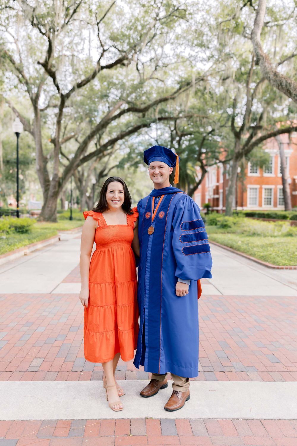 Two people pose together on a brick path with Spanish moss draped trees and a red brick building in the background.