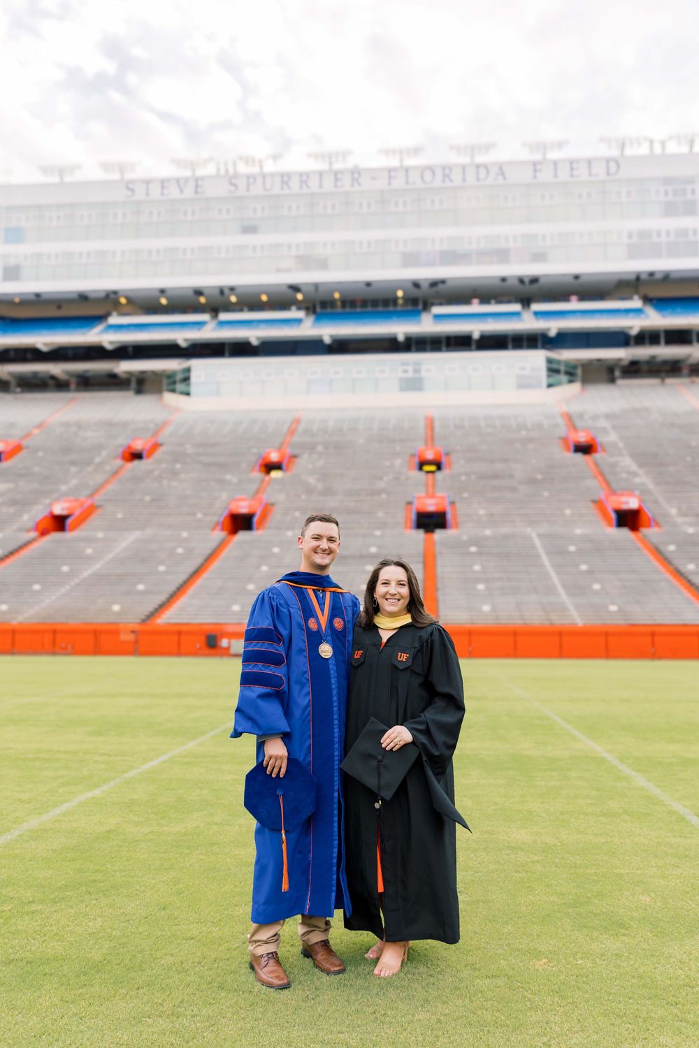 Two graduates in academic regalia pose on a football field with stadium seating and orange pylons in the background.