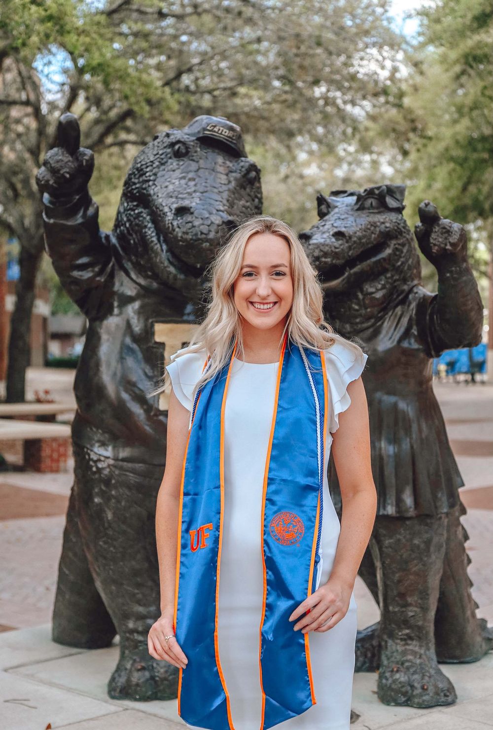 Graduate in a blue stole poses with cheerful bronze statues on campus plaza.