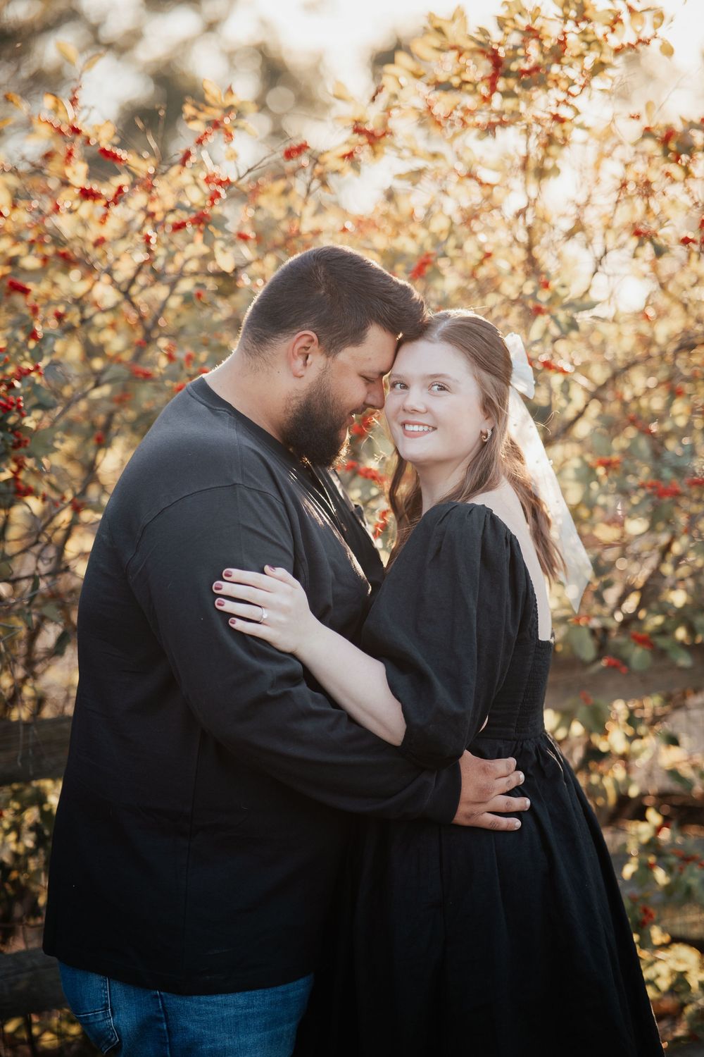 A couple in matching black outfits share an intimate moment among fall foliage with red berries in golden sunlight.