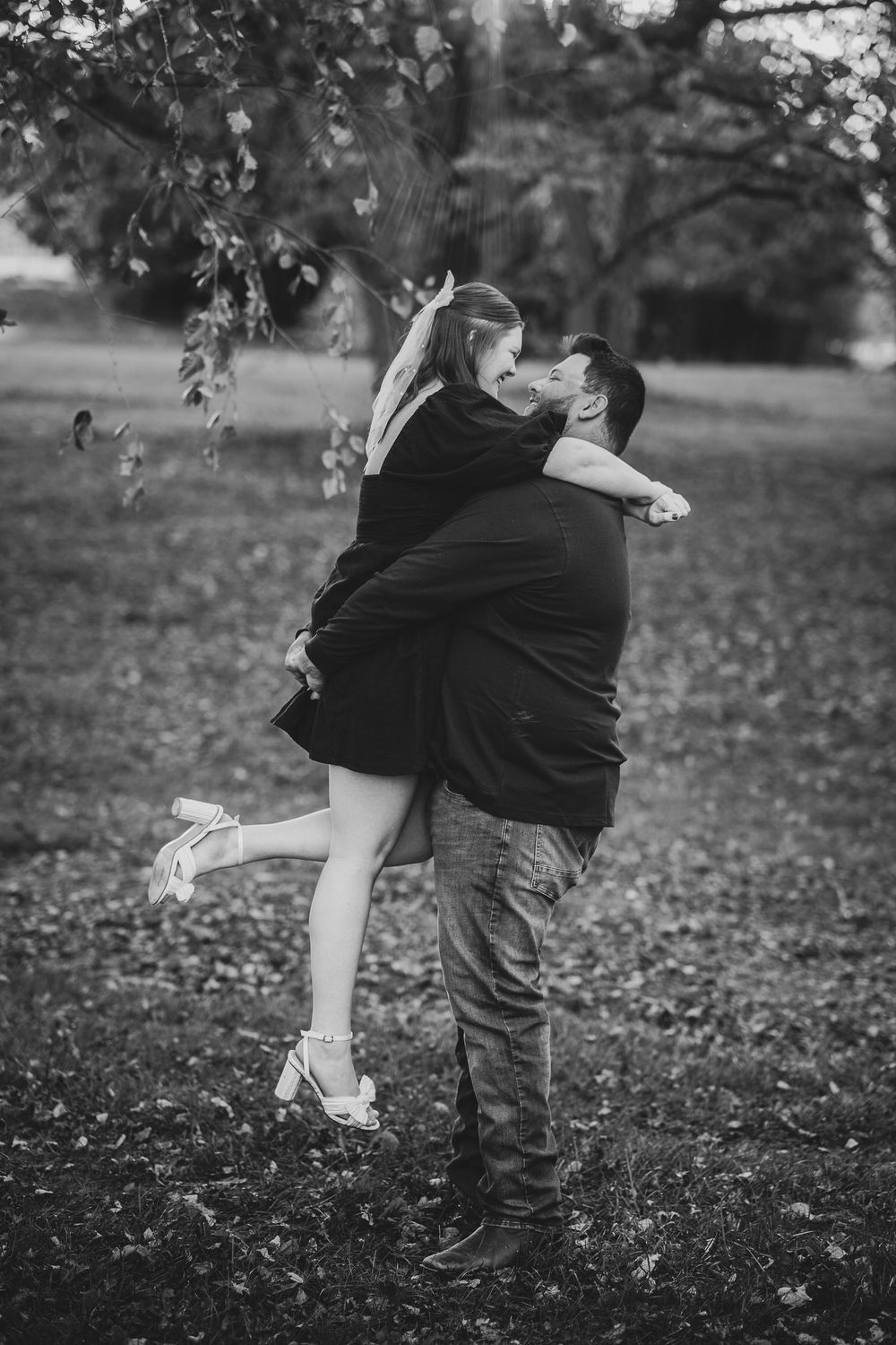 A romantic black and white photo of a couple embracing outdoors under trees with fallen leaves on the ground.