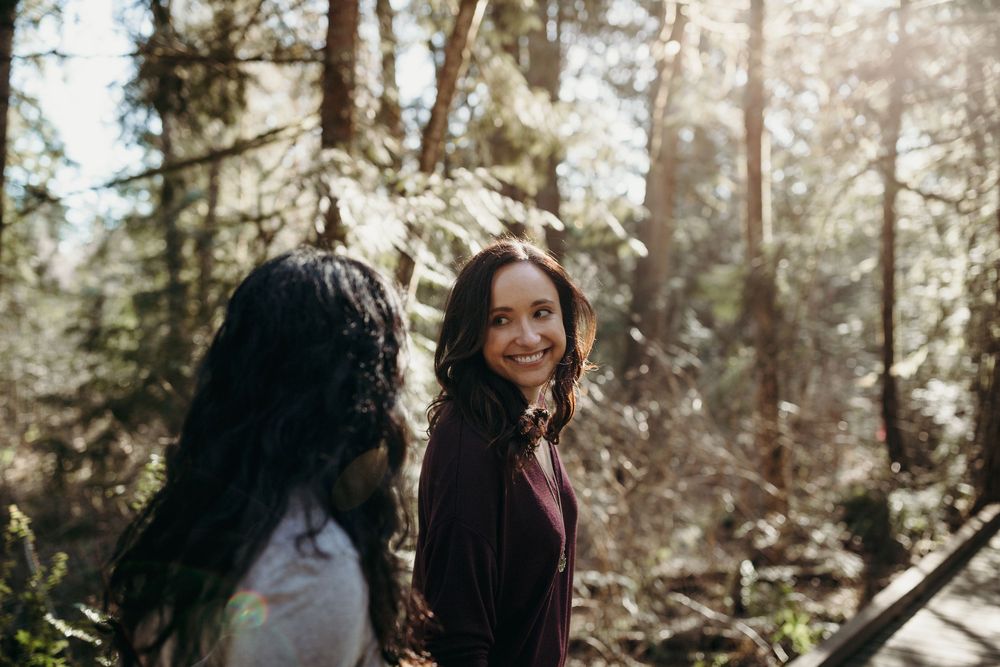 Two people walking through sunlit forest surrounded by tall evergreen trees during golden hour.