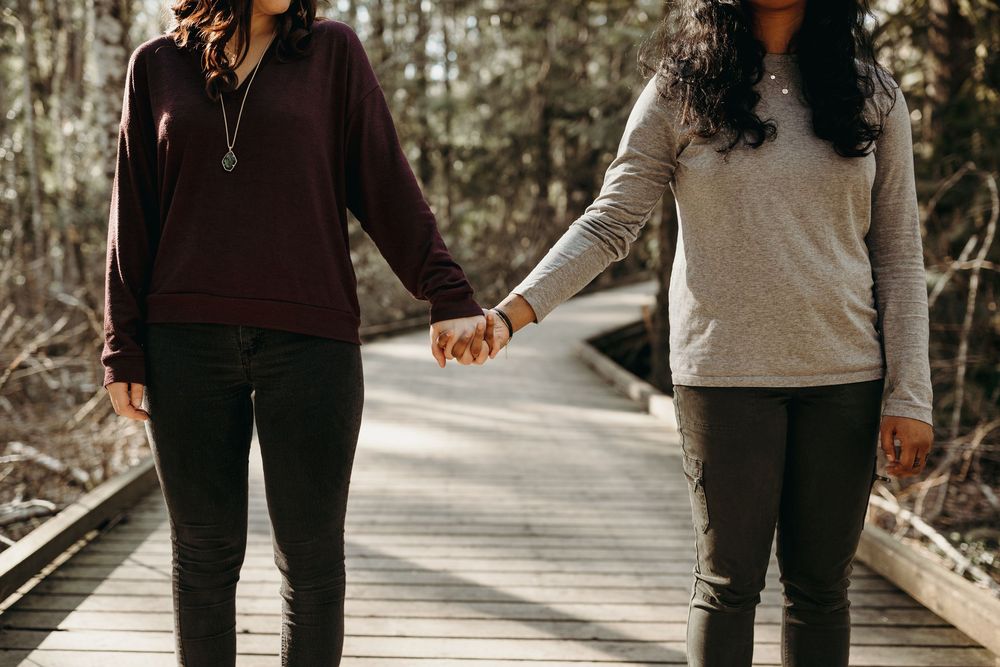 Two people hold hands while walking together on a wooden boardwalk surrounded by trees in autumn.