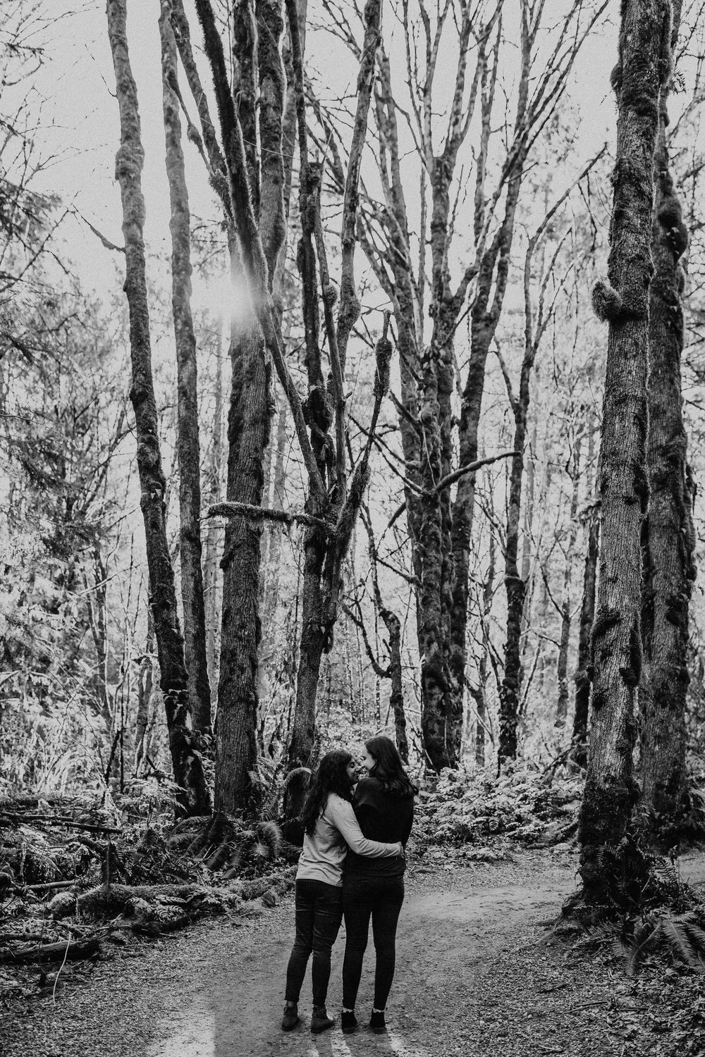 A couple embraces while walking down a serene forest path surrounded by towering trees in black and white.