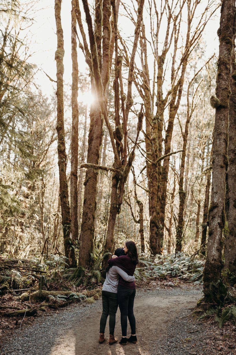 A couple embraces on a sunlit forest trail surrounded by tall trees on a misty morning.