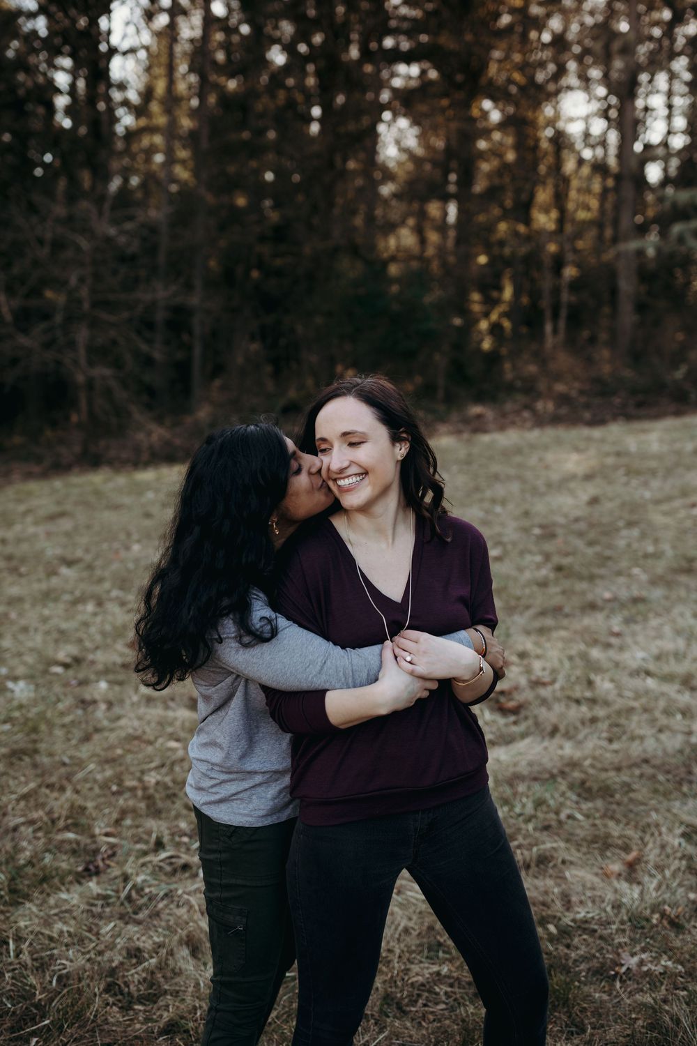 A couple embraces lovingly while standing in a grassy field during golden hour with forest in the background.