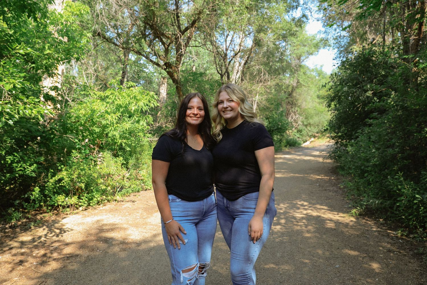 Two friends in matching black shirts and jeans pose on a sunlit nature trail.