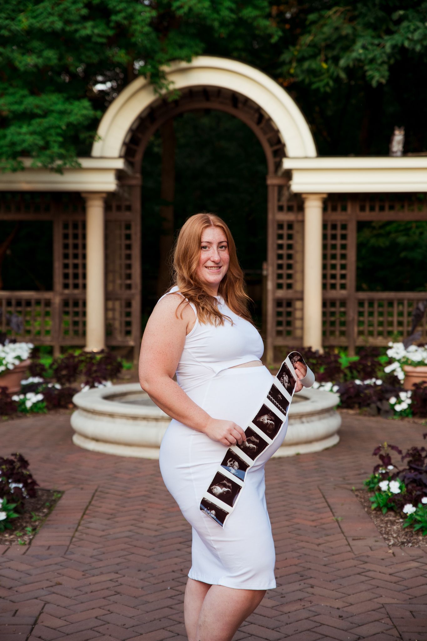 Maternity photo of woman in white dress at garden archway.