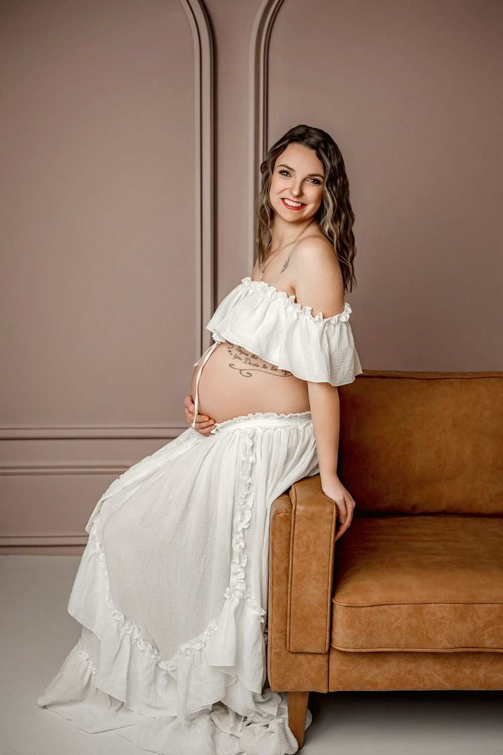 Maternity photoshoot featuring elegant white dress poses on brown leather couch against neutral wall backdrop.