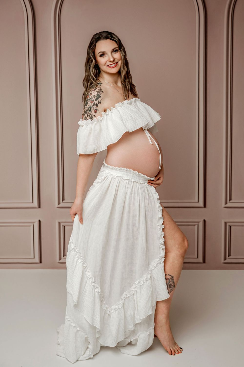 A maternity photo series showing a pregnant woman in a flowing white dress against a brown paneled wall.
