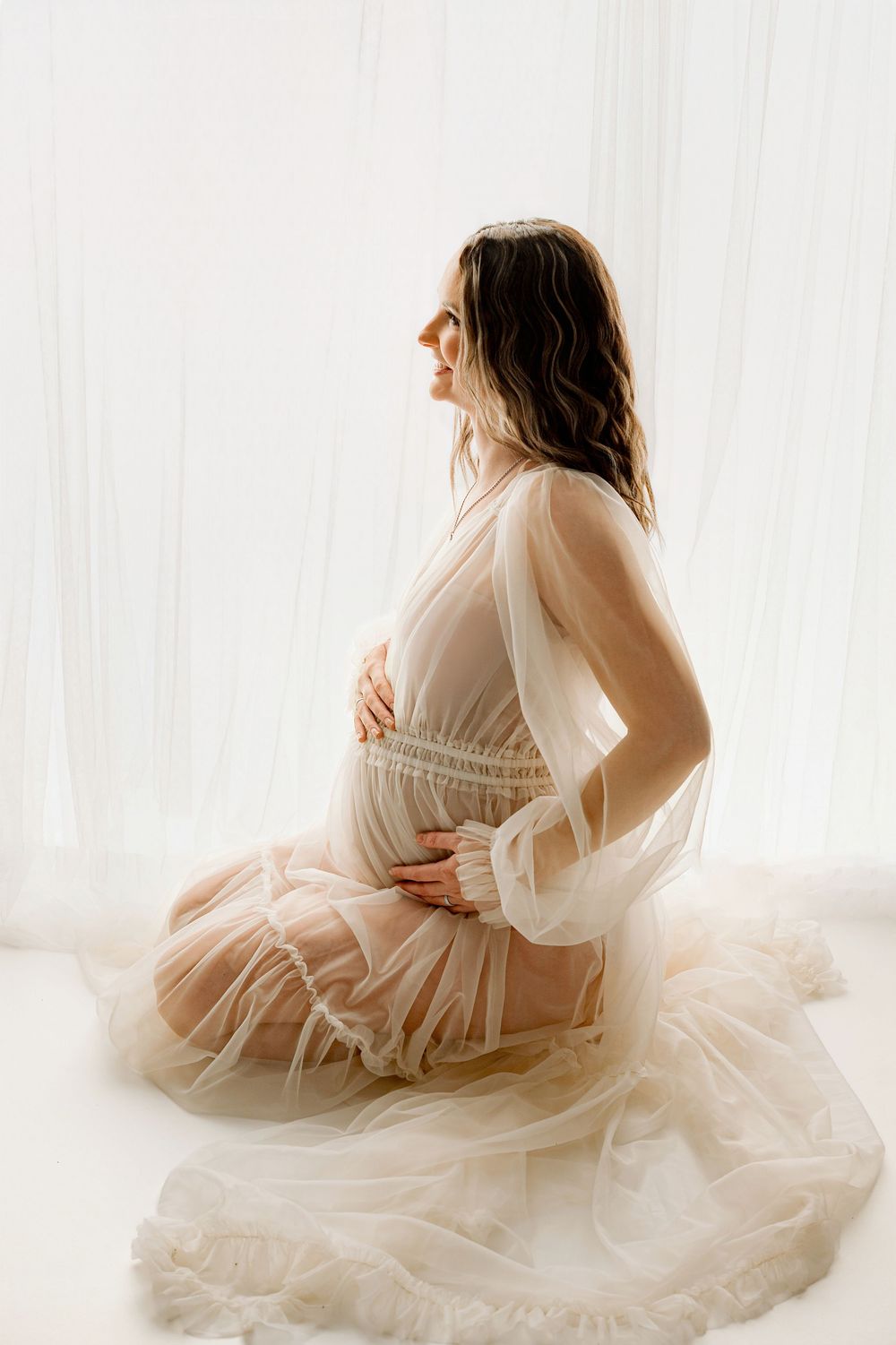 Ethereal maternity photos in white flowing dress against soft window light creating dreamy silhouettes.