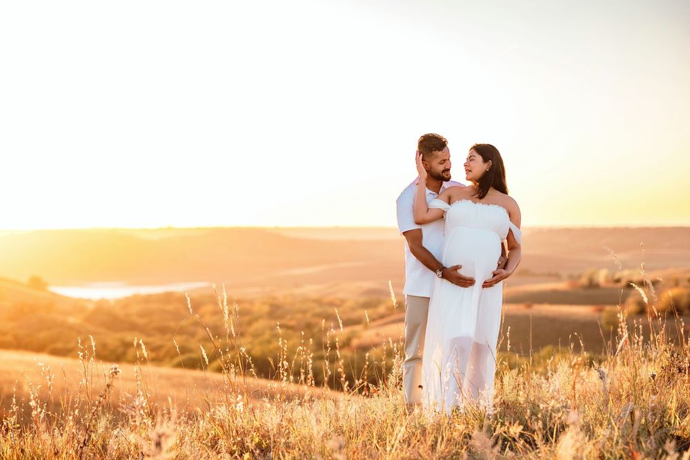 A couple in white outfits embraces in a golden wheat field at sunset with rolling hills in the background.