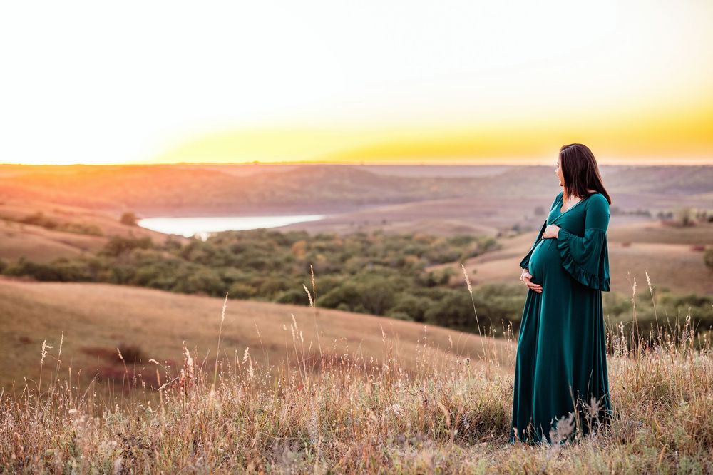 A person in a teal dress stands in a golden field at sunset with rolling hills in the background.