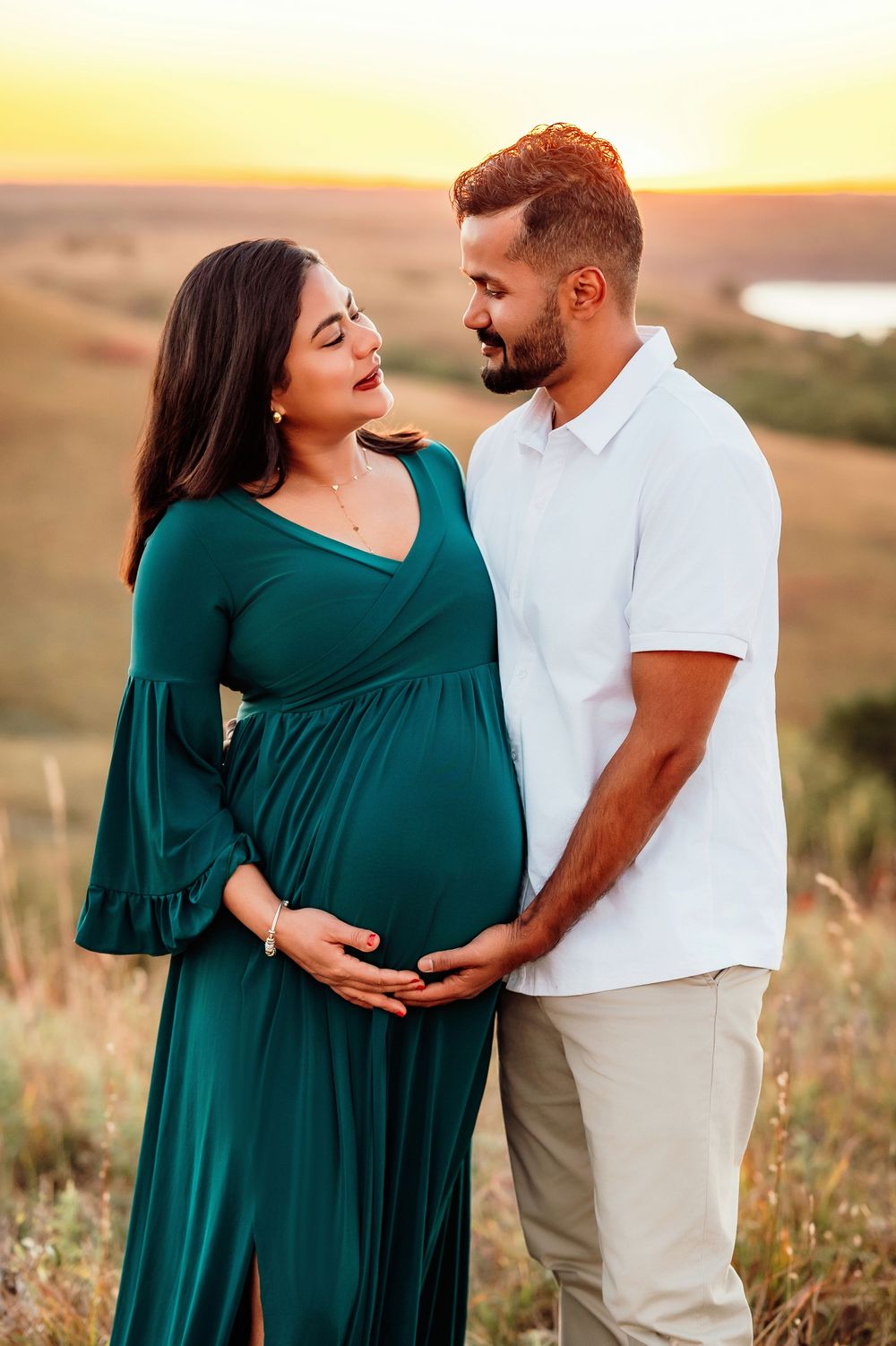 A couple shares an intimate moment at sunset, with the woman wearing an emerald green maternity dress in a scenic field.