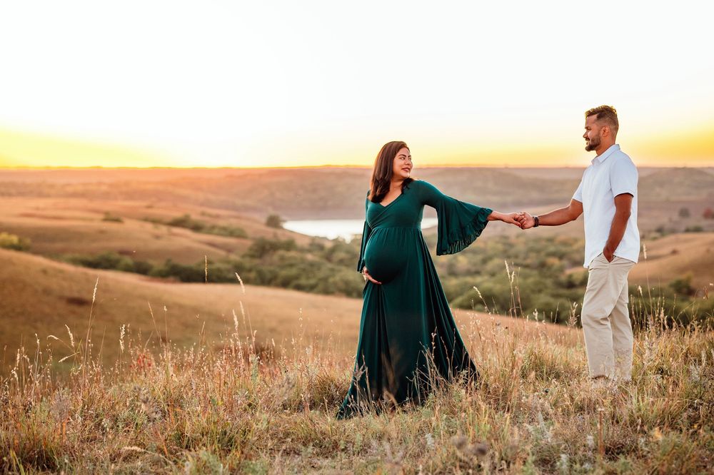 A couple holds hands on a hilltop during golden hour sunset, with rolling hills and grasslands in the background.