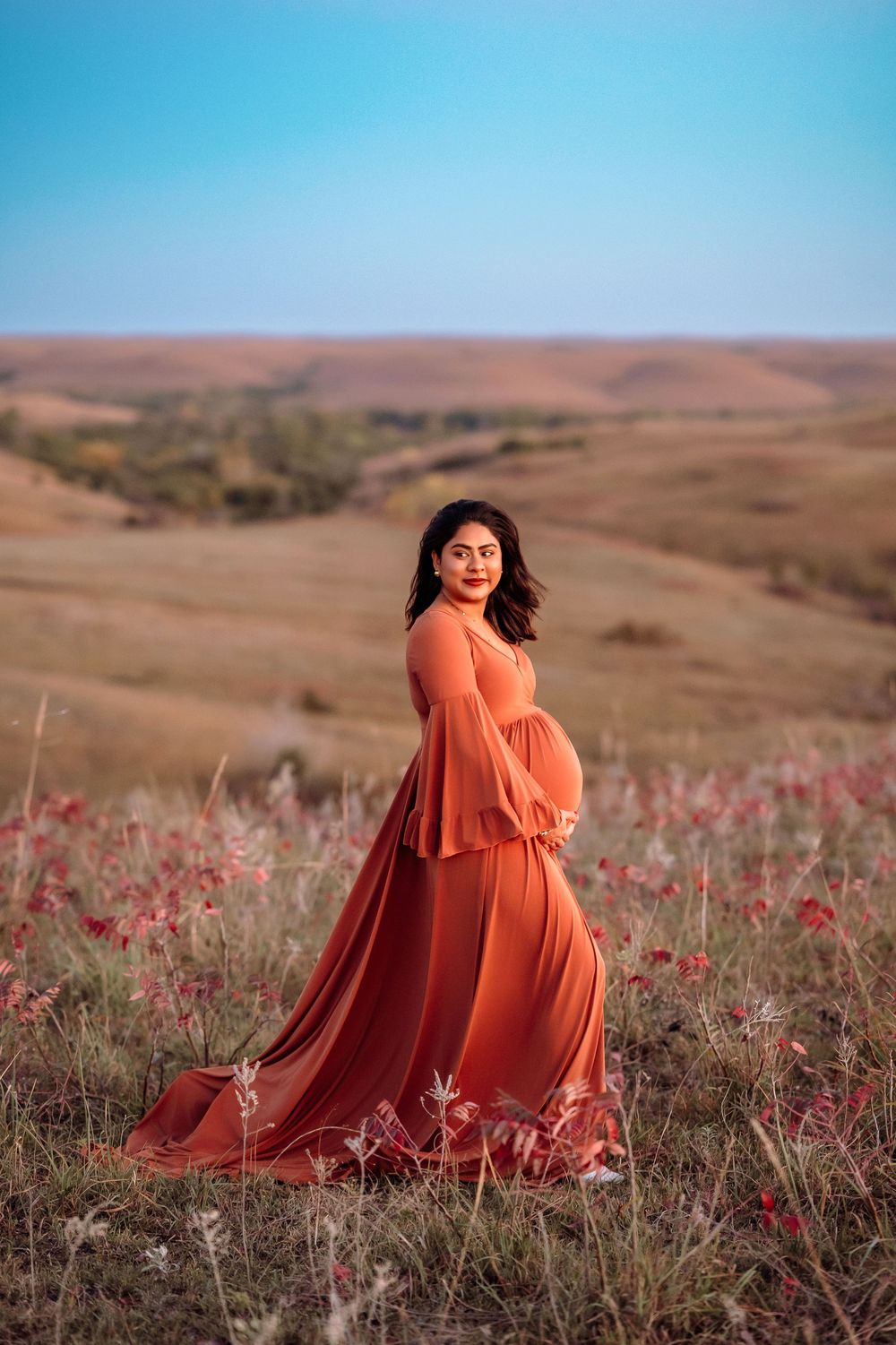 A flowing orange dress twirls gracefully across a desert landscape with scattered wildflowers at sunset.