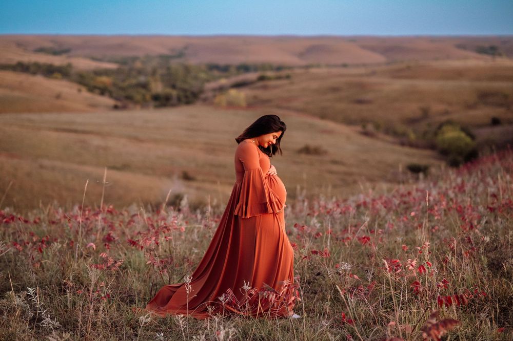 A figure in a flowing orange dress walks through a field of vibrant red wildflowers against rolling hills and blue sky.