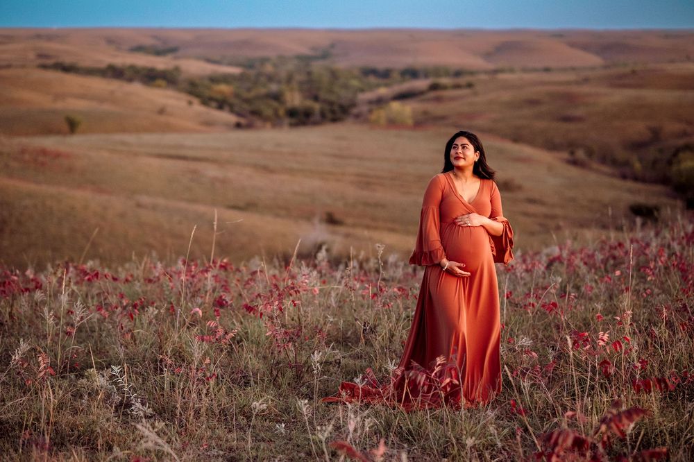 A bright orange dress stands out against rolling brown hills dotted with vibrant wildflowers at sunset.