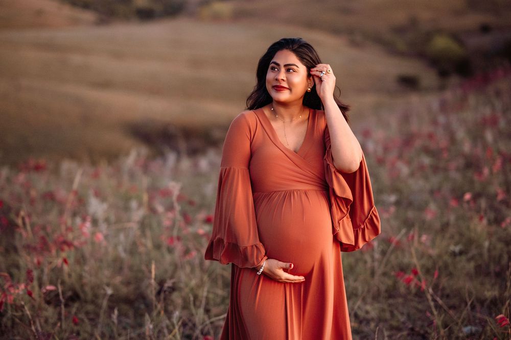 Expecting mother in flowing burnt orange dress poses in field of wildflowers at sunset.