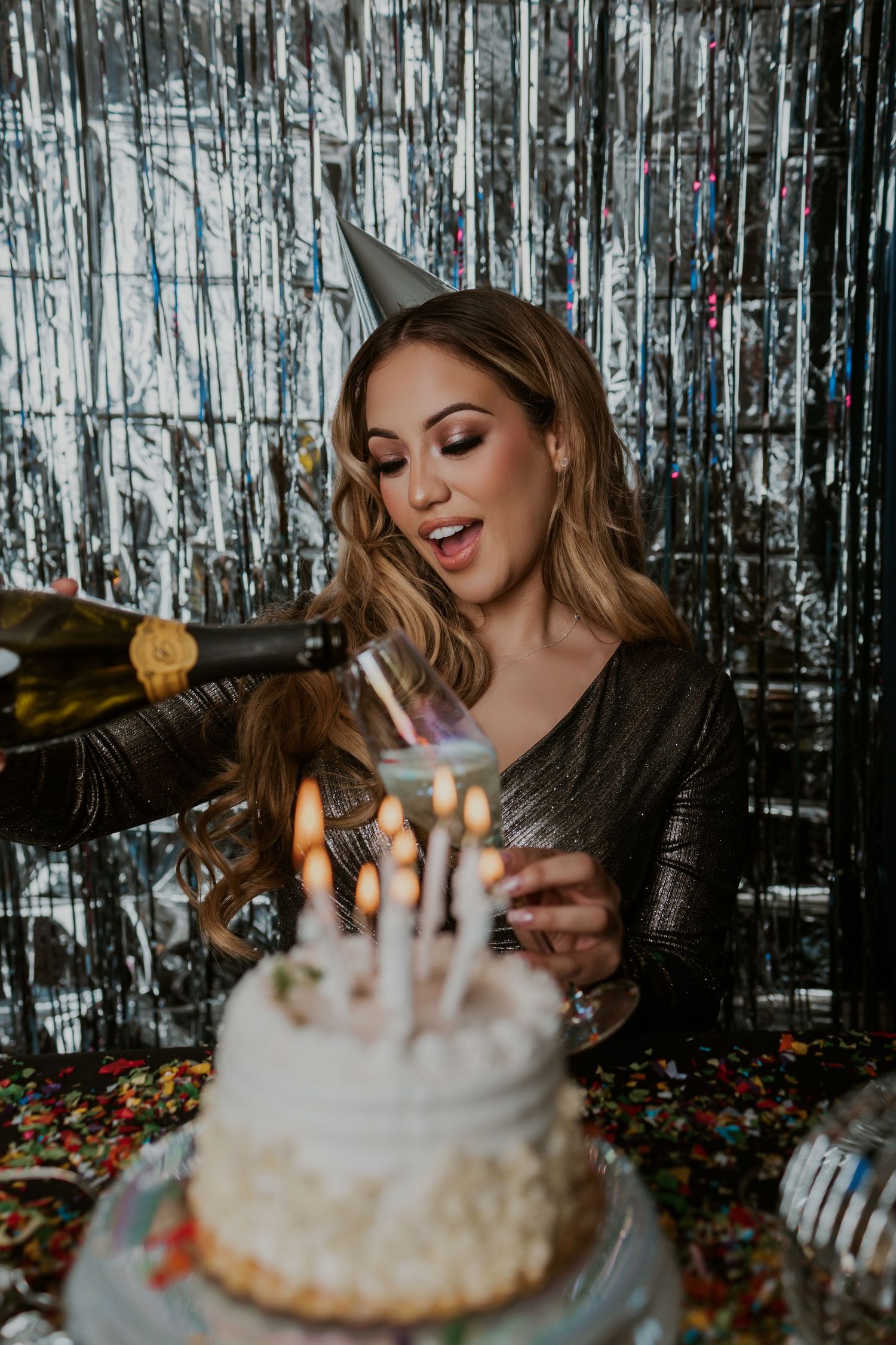 Woman celebrating with birthday cake and champagne against silver tinsel backdrop.